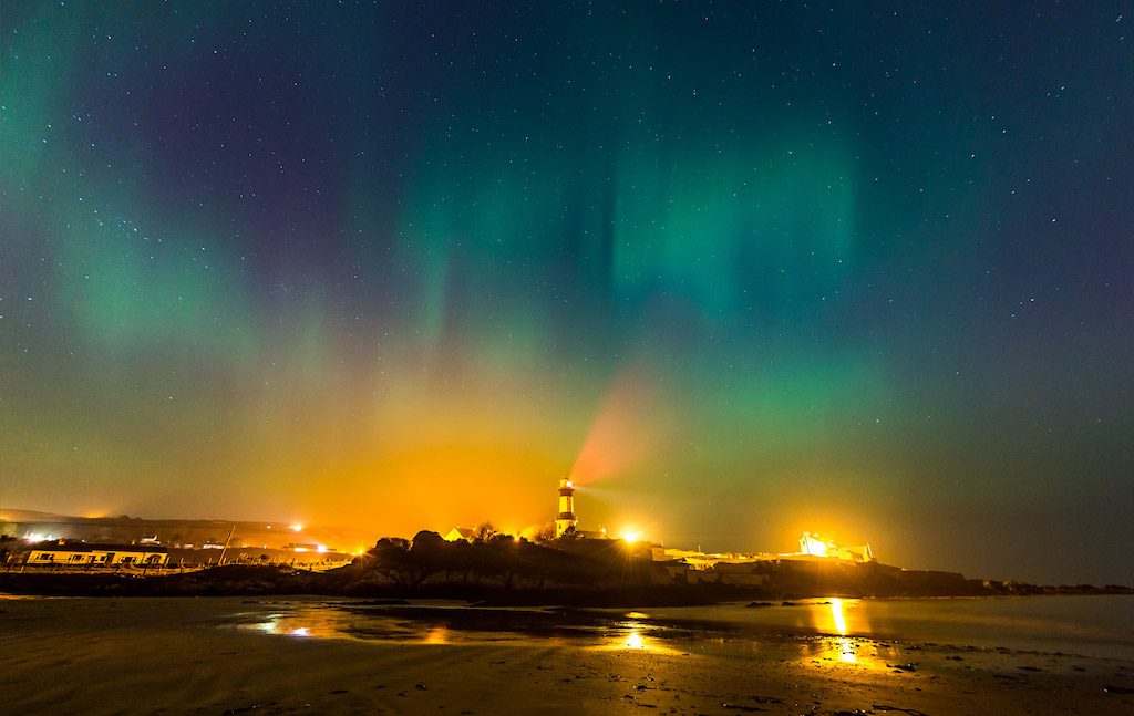 Northern lights glow above Stroove Lighthouse with golden reflections on the shore in County Donegal.