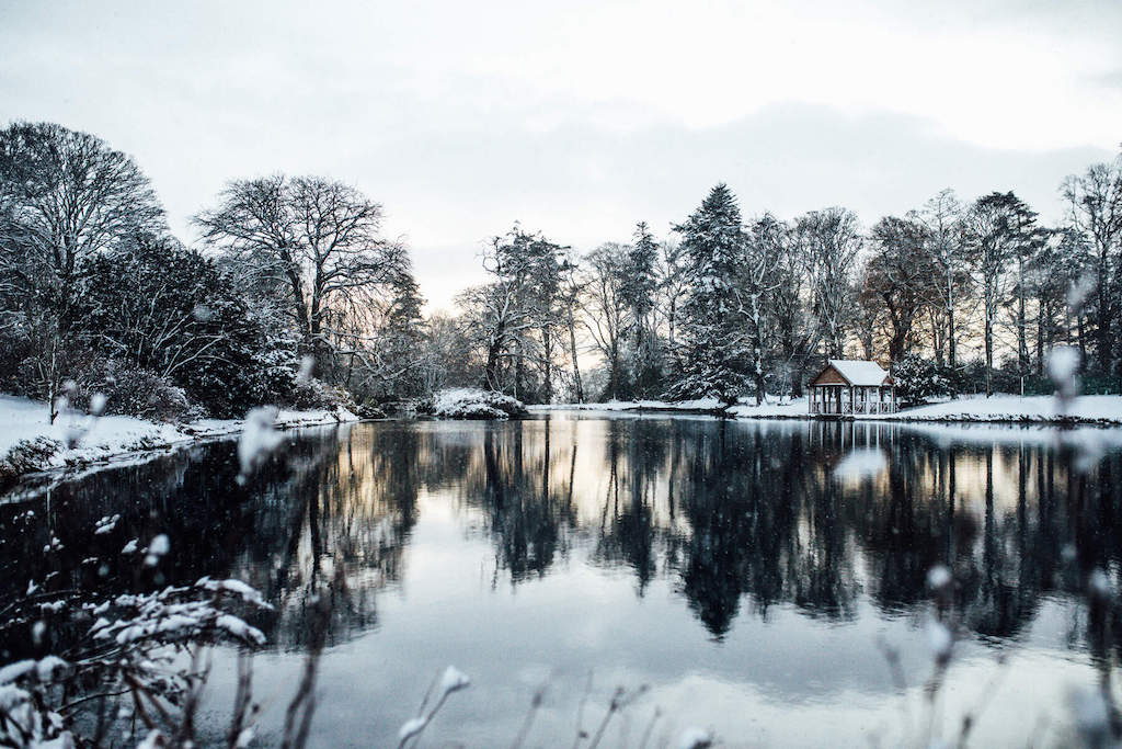 Snow-covered trees reflected in a still lake with a small wooden hut at Montalto Estate, County Down.